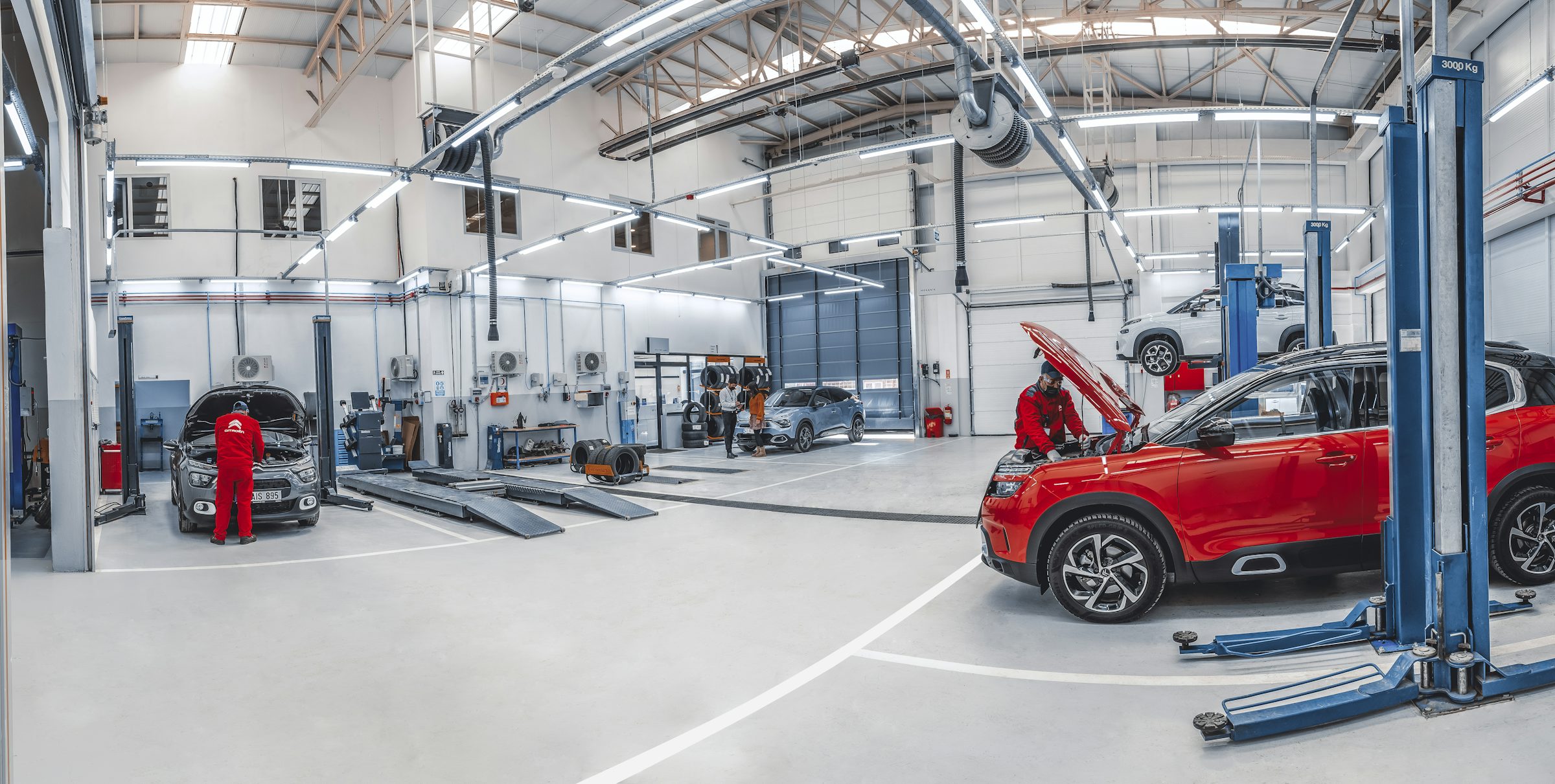 Vehicles on lifts in a bright inspection workshop, with a technician checking a car on the bay